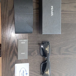 Svarta solglasögon från Prada - Snygga svarta solglasögon från Prada med breda skalmar och tydlig logga i guld på sidorna. Glasögonen har en modern, kantig form och levereras med originalfodral, putsduk och äkthetskort. Perfekta för dig som vill ha en lyxig och stilren accessoar.