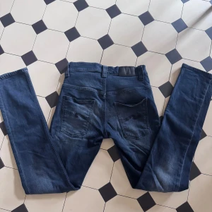 Blå jeans från Nudie Jeans W28 L32 - Säljer ett par klassiska blå jeans från Nudie Jeans i modellen W28 L32. Jeansen har raka ben, fem fickor och snygga slitningar på framsidan. Tillverkade i mjukt denimtyg med Nudie-logga på bakfickorna. Perfekta för en avslappnad och stilren look.