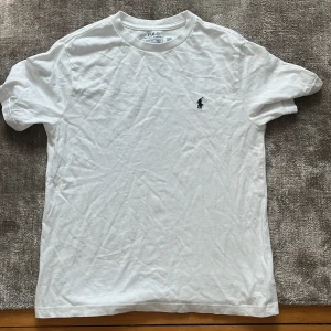 Vit t-shirt från Polo Ralph Lauren - Klassisk vit t-shirt från Polo Ralph Lauren med broderad marinblå logga på bröstet. T-shirten är i mjuk bomull, har rund hals och korta ärmar. Perfekt basic-plagg med snygg passform och stilren look.