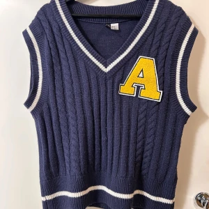 Mörkblå stickad väst med A-patch - Collegeinspirerad mörkblå stickad väst med vita ränder vid hals, ärmöppningar och nederkant. Västen har en stor gul bokstav A på bröstet och v-ringad hals. Snygg kabelstickad struktur som ger en sportig och trendig look.