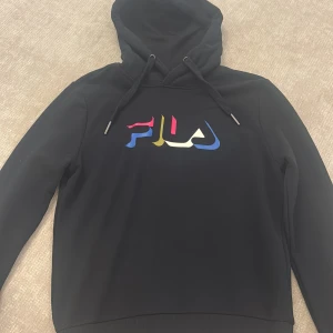 Svart hoodie från FILA med färgglad logga - Svart hoodie från FILA med stor, färgglad logga på bröstet i blått, rött, gult och vitt. Tröjan har huva med snörning och långa ärmar. Perfekt för vintern. .