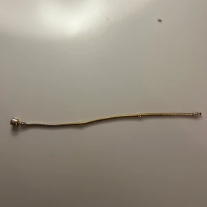 Pandora guldarmband med lås - Snyggt armband från Pandora i guldfärgat material med klassiskt ormkedjemönster. Låset är cylinderformat och har Pandora-loggan ingraverad. Perfekt för att samla dina favoritberlocker och skapa en personlig stil.