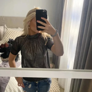 Glittrig svart blus med hög hals - Säljer en svart glittrig blus med hög hals och korta ärmar. Blusen är transparent med en snygg glansig finish. Perfekt för dig som vill sticka ut med en cool och trendig look. Passar perfekt till fest och nyår! 🩷🩷 Den är i storlek xs från HM! Ops lånade bilder!