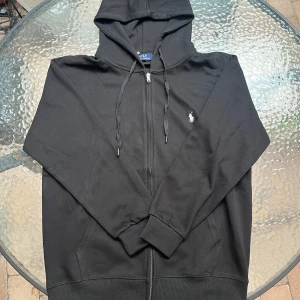 Svart hoodie från Polo Ralph Lauren - Svart hoodie från Polo Ralph Lauren med dragkedja, huva med snörning och klassisk vit broderad logga på bröstet. Tillverkad i mjuk bomull och har ribbade muddar samt fickor framtill. Perfekt för en chill och stilren look. Det är en XL men den är mycket liten i storleken.