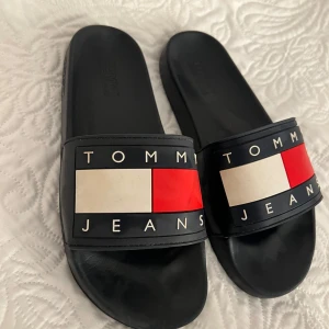Svarta slides från Tommy Jeans - Svarta sandaler från Tommy Jeans med klassisk logga i rött, vitt och blått framtill. Snygg och enkel design med bred rem och platt sula, perfekta för sommaren. Tillverkade i syntetmaterial och har öppen tå för en chill vibe.
