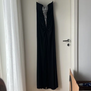 Svart långklänning med strass KDK London - Supersnygg svart långklänning från KDK London med axelbandslös design och glittrande strassdetaljer framtill. Klänningen har draperad överdel och markerad v-ringning, perfekt för bal eller fest. Materialet känns mjukt och följsamt.