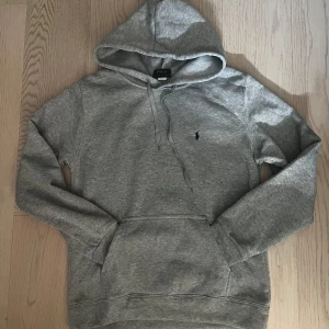 Grå hoodie från Polo Ralph Lauren - Snygg grå hoodie från Polo Ralph Lauren med klassisk broderad logga på bröstet. Tröjan har huva med snörning, känguruficka och långa ärmar. Perfekt för chill dagar och enkel att matcha med jeans eller joggers.