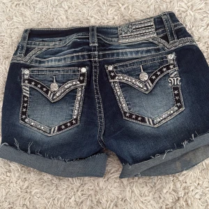 Miss Me jeansshorts med stjärndetaljer - Snygga mörkblå jeansshorts från Miss Me som ja klippt själv med coola stjärndetaljer, paljetter och broderier på fickorna. Shorts med råa kanter och uppvikta benslut, silverfärgade knappar och kontrastsömmar. Perfekta för dig som vill sticka ut med en unik och edgy stil. Alla paljetter och pärlor sitter kvar! Den enda defekten är en blekningsfläck (därför jag klippte de) men syns ej när man viker upp de o man kan sy de också. 