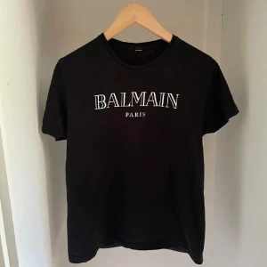 Svart Balmain Paris t-shirt - Svart t-shirt från Balmain Paris med vit logotyp framtill. Klassisk rund halsringning och korta ärmar. Tillverkad i mjuk bomull för en skön känsla och clean look. Perfekt för dig som gillar stilrena statement-plagg.
