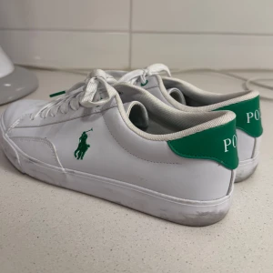 Vita sneakers från Polo Ralph Lauren - Fräscha vita sneakers från Polo Ralph Lauren med gröna detaljer och logga på sidan samt hälen. Skorna har klassisk snörning, rund tå och platt sula. Ovandelen är i syntetmaterial och insidan har grön innersula med Polo-logga. Perfekta för en clean och sportig look. Använd 3 gånger, defekterna syns på sista bilderna 