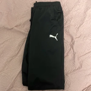 Svarta träningsbyxor från Puma - Mycket sköna att ha på sig dom och är svåra att hitta. Svarta träningsbyxor från Puma med vit logga på låret. Byxorna har elastisk midja och dragkedja vid benslut för extra komfort. Tillverkade i mjukt syntetmaterial som passar perfekt för sport och chill. Snygg och enkel design med normal passform.