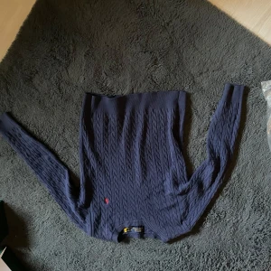 Marinblå kabelstickad tröja - Snygg marinblå kabelstickad tröja från Polo Ralph Lauren med klassisk röd logga broderad på bröstet. Tröjan har rund hals och långa ärmar, perfekt för dig som gillar stilren och tidlös design.