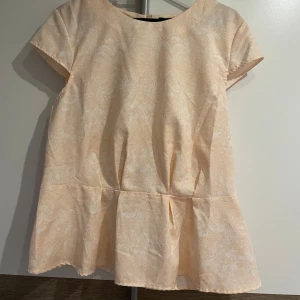 Ljus rosa blus  - Supersöt  ljus rosa blus med subtilt mönster och volang nertill. Rund halsringning och korta ärmar ger en clean och fräsch look. 