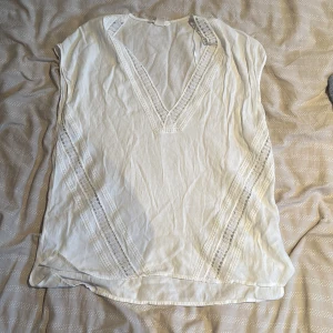 Vit blus med spetsdetaljer H&M - Vit blus från H&M i storlek 40 med v-ringning och fina spetsdetaljer längs halsringning och sidor. Blusen är ärmlös och har en luftig känsla, perfekt för varmare dagar. Materialet är mjukt och lätt, vilket gör den superbekväm att bära.