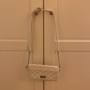 Vit quiltad axelväska från Justice - Snygg vit axelväska från Justice med quiltat mönster och små silvriga nitar. Väskan har en kombinerad axelrem i kedja och läderimitation, samt ett metallmärke framtill. Perfekt storlek för mobil och småprylar. Är Sundling välanvänd, men fortfarande väldigt fin. Därav billigt pris