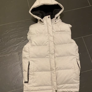 Vit rutig dunjacka från Everest - Vit dunjacka från Everest med rutigt mönster, huva och dragkedja. Jackan är vadderad och har praktiska fickor med dragkedja samt avtagbar huva. Perfekt för kalla dagar när du vill hålla stilen och värmen.