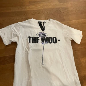 Vit THE WOO t-shirt med rostryck - Säljer en vit t-shirt från THE WOO i 100% bomull, tillverkad i USA. Framsidan har ett stort svart tryck med texten 'THE WOO-' och en stiliserad ros i lila och svart. Baksidan har ett unikt mönster med rosor i grått och svart. Klassisk passform och korta ärmar.