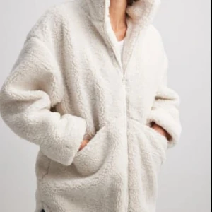 Vit teddyjacka med dragkedja - Mysig och oversized vit teddyjacka med hög krage och praktiska fickor framtill. Jackan har en grov, fluffig struktur och stängs med dragkedja. Perfekt för kalla dagar när du vill ha både stil och komfort. Overzied i modellen.