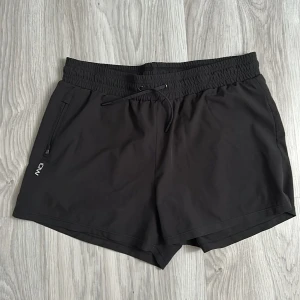 Svarta träningsshorts från ICANIWILL - Svarta träningsshorts från ICANIWILL med elastisk midja och snörning. Shortsen har dragkedjeficka på sidan och är tillverkade i ett lätt syntetmaterial som passar perfekt för gym eller löpning. Snygg minimalistisk look med logga på benet.