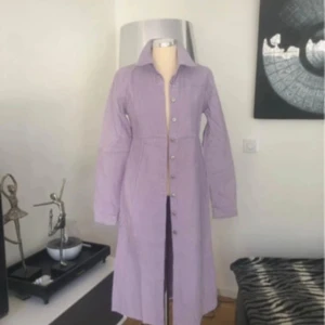 Ljuslila långklänning i bomull - Långklänning i ljuslila bomull med klassisk krage och knappar hela vägen framtill. Klänningen har långa ärmar med manschett och markerad midja för en snygg siluett. Perfekt för dig som gillar stilrena och bekväma plagg. Kan användas som kappa/kimono