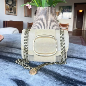 Beige axelväska från See by Chloé - Supersnygg beige axelväska från See by Chloé med guldfärgad kedja och stor dekorativ spänne framtill. Väskan är gjord i en mix av skinn och mocka, och har en exklusiv känsla med detaljer i guld. Perfekt storlek för det viktigaste och ger en lyxig touch till din outfit. Den har dock använts under nått år nu så är inte i nyskick längre men det är inget som syns på längre håll :)
