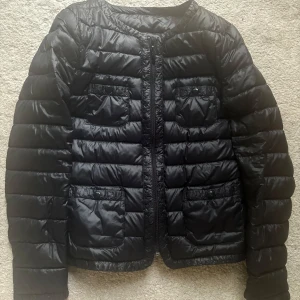 Svart quiltad pufferjacka - Säljer en svart pufferjacka med quiltad design och rund halsringning. Jackan har fyra fickor framtill med knappstängning och dragkedja i mitten. Perfekt för kyliga dagar och enkel att styla till olika outfits.