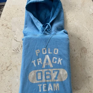 Blå hoodie från Polo Ralph Lauren - Snygg blå hoodie från Polo Ralph Lauren med tryck 'Polo Track 067 Team' framtill. Tröjan har huva med snörning och en klassisk känguruficka. Mjuk bomullskvalitet och cool sportig vibe, perfekt för dig som gillar streetwear.