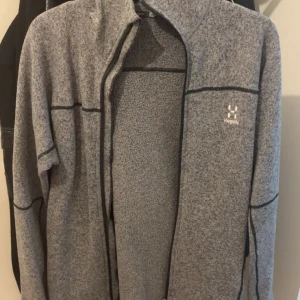 Grå fleece hoodie från Haglöfs - Säljer en gråmelerad fleece hoodie från Haglöfs med svarta detaljer och hel dragkedja. Tröjan har hög krage, långa ärmar och en skön huva. Perfekt för kyliga dagar och outdoor-häng. Klassisk logga på bröstet och mjukt material.