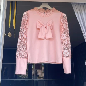 Rosa blus med spetsärmar och rosett - Supersöt ljusrosa blus med stora spetsärmar och en maffig rosett framtill. Ärmarna är i genombruten spets med blommönster och blusen har volangkrage. Perfekt för dig som gillar gulliga detaljer och romantisk stil.