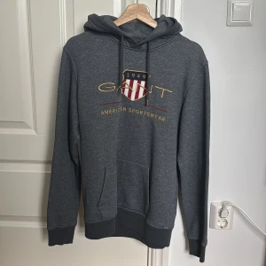 Mörkgrå hoodie - GANT - Säljer denna mörkgrå hoodie från GANT med broderat emblem och text på bröstet. Har använts max 5 gånger. Använder inte den då den är lite stor för mig. Inga skador, smuts eller något liknanden alls. Vill du ha bilder på något specifikt är det bara att höra av dig då!