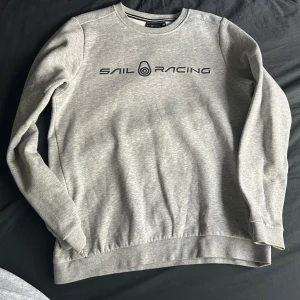 Grå Sail Racing sweatshirt - Snygg grå sweatshirt från Sail Racing med svart logotyp tryckt på bröstet och en liten logga på ryggen. Tröjan har rund hals, långa ärmar och ribbade muddar. Perfekt för chill dagar och enkel att matcha med jeans eller joggers.
