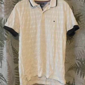 Vit piké från Tommy Hilfiger M - Vit pikétröja från Tommy Hilfiger i regular fit. Klassisk krage med mörkblå och röda detaljer, korta ärmar med mörkblå muddar och diskret logga på bröstet. Tillverkad i 100% bomull för en skön känsla.
