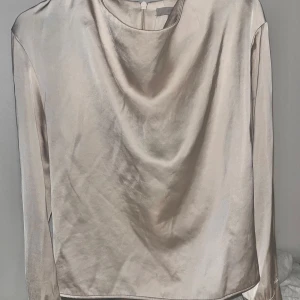 Champagnefärgad satintopp med drapering - Snygg långärmad topp i champagnefärgad satin med draperad halsringning. Tidlös och elegant look med mjukt, glansigt material som faller fint. Perfekt för dig som gillar stilrena plagg med en twist.