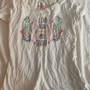 Vit t-shirt med tryck från Pepperts - Vit t-shirt från Pepperts med coolt tryck framtill: färgglada fjädrar, geometriska former i rosa, orange och grönt samt texten 'Live now and enjoy'. Rund halsringning och korta ärmar. Skön bomullskvalitet, perfekt till sommaren. Pris kan diskuteras.