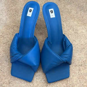 Blå klackade sandaler från Ginatricot - Snygga blå sandaler från Ginatricot med fyrkantig tå och vadderad, tvistad rem över foten. Skorna har klack och är tillverkade i ett mjukt syntetmaterial som ger en cool statement-look. Perfekta för dig som vill sticka ut med färg och form.