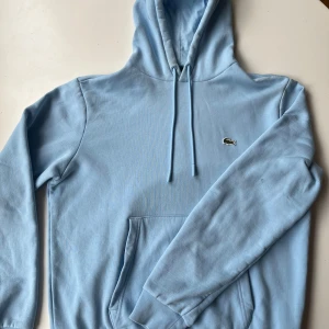 Ljusblå hoodie från Lacoste - Snygg ljusblå hoodie från Lacoste med klassisk krokodillogga på bröstet. Tröjan har huva med snörning, känguruficka och ribbade muddar. Perfekt för dig som gillar stilrena och bekväma plagg. Materialet är mjuk bomull som känns skönt mot huden.