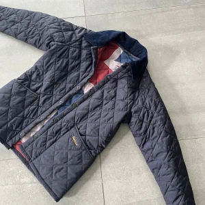 Svart quiltad jacka från Barbour - Quiltad svart jacka från Barbour med klassisk krage i manchester, tryckknappar och två stora fickor framtill. Jackan har Barbour-logga broderad på fickan och ett snyggt rutigt innerfoder. Perfekt för dig som gillar stilren och tidlös look.