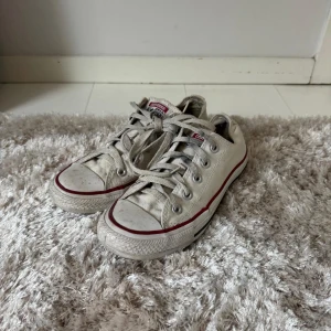 Vita Converse All Star sneakers - Klassiska vita Converse All Star sneakers med lågt skaft, röd och blå rand runt sulan och snörning framtill. Skorna är i canvas med rund tå och platt sula. Perfekta för en avslappnad och cool stil.