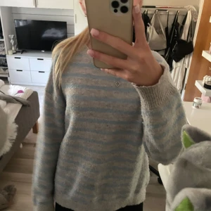 Randig stickad tröja Gina Tricot - Mysig stickad tröja från Gina Tricot i ljusgrått och ljusblått randigt mönster. Tröjan har rund halsringning och långa ärmar, perfekt för lager-på-lager under kyliga dagar. Skön och avslappnad passform som funkar till allt.🩵