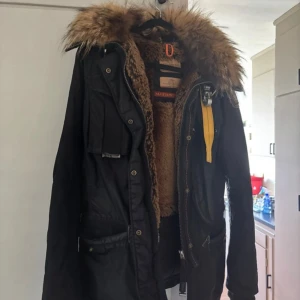 Svart Parajumpers parkas med päls - Säljer en svart parkas från Parajumpers med fluffig brun päls runt huvan och varm dunfyllning. Jackan har flera fickor, dragkedja, knappar och den ikoniska gula remmen vid kragen. Insidan är fodrad med mjuk teddy och jackan är perfekt för kalla dagar.  i fint skick! Men lite slitage på vissa skinndetsljer , det jag tagit bild på. Inte använd supermycket, men slitaget uppkom tyvär vid en rengöring av ja