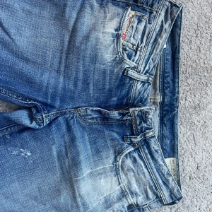 Diesel bootcut jeans  - Säljer ett par blå bootcut jeans från Disel med slitna detaljer och snygga bakfickor med V-formad söm. Jeansen har klassisk femficksdesign, dragkedja och knapp, samt tydliga slitningar för en cool look. Perfekta för dig som gillar avslappnad stil.