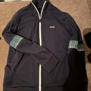 Mörkblå ziptröja från BOSS - Snygg mörkblå tröja från BOSS med vit dragkedja och logga på bröstet. Ärmarna har tryckt BOSS-mönster i vitt och ljusgrönt. Tröjan har hög krage och ribbade muddar. Perfekt för en stilren och sportig look.