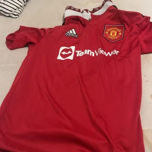 Manchester United Adidas fotbollströja - Röd Manchester United fotbollströja från Adidas med klubbmärke på bröstet och TeamViewer-logga. Klassisk design med vita detaljer vid kragen och Adidas-logga. Tillverkad i lätt och ventilerande material, perfekt för match eller träning.