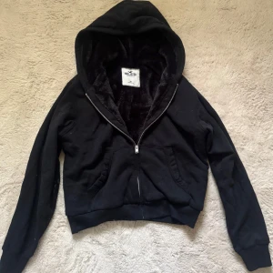 Svart hoodie från Hollister med dragkedja - Svart hoodie från Hollister med mjukt foder och huva. Tröjan har dragkedja framtill, två fickor och långa ärmar. Perfekt för kyliga dagar och har en skön, lite loose passform. 