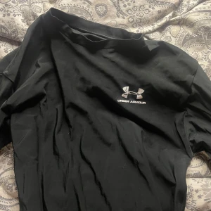 Svart Under Armour t-shirt - Svart t-shirt från Under Armour med klassisk logga i vitt på bröstet. T-shirten har korta ärmar och rund hals. Materialet känns som mjuk bomull, perfekt för träning eller chill. På baksidan finns ett ovalt tryck med färgstänk i rött, vitt och grått.