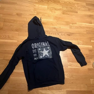Svart hoodie med tryck och huva - Säljer en svart hoodie med stor vit tryckt text 'ORIGINAL FIT EST. 2024' och en stjärna med mindre stjärnor på bröstet. Tröjan har huva med snörning och en klassisk magficka. Perfekt för dig som gillar streetstyle och vill ha något bekvämt och snyggt.