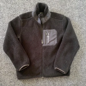 Svart fleecejacka från Sail Racing - Mysig svart fleecejacka från Sail Racing med hög krage och dragkedja framtill. Jackan har en praktisk bröstficka med dragkedja och detaljer i grått. Perfekt för kyliga dagar och enkel att slänga på över en hoodie.