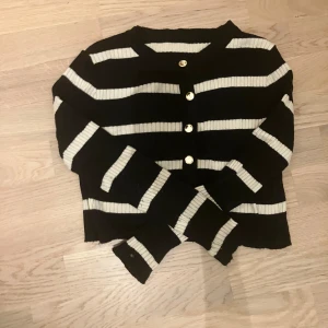 Svartvit randig kofta med guldknappar - Säljer en svart och vit randig kofta med ribbad struktur och blanka guldknappar framtill. Koftan har långärmade armar och är croppad i modellen. Perfekt för dig som gillar klassiska mönster med en twist. Superenkel att matcha med jeans eller kjol.