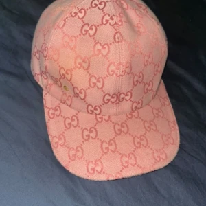Rosa Gucci keps med GG-mönster - KOMMER MED SJÄLVA BOXEN!!!!!!Säljer en snygg rosa keps från Gucci med ikoniskt GG-mönster över hela kepsen. Kepsen har justerbart spänne i rosa läder baktill och är tillverkad i bomull. Perfekt för dig som vill sticka ut med en lyxig och trendig accessoar.