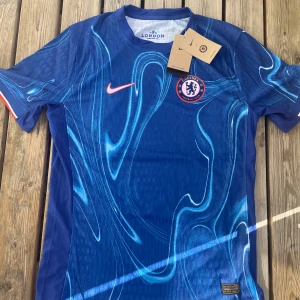 Chelsea FC blå matchtröja Nike - Chelsea FCs officiella matchtröja från Nike i blått med unikt abstrakt mönster i ljusare blå toner. Kortärmad modell med klubbmärke på bröstet och röd Nike-logga. Tillverkad i lätt och ventilerande material, perfekt för fotboll eller supporterliv.